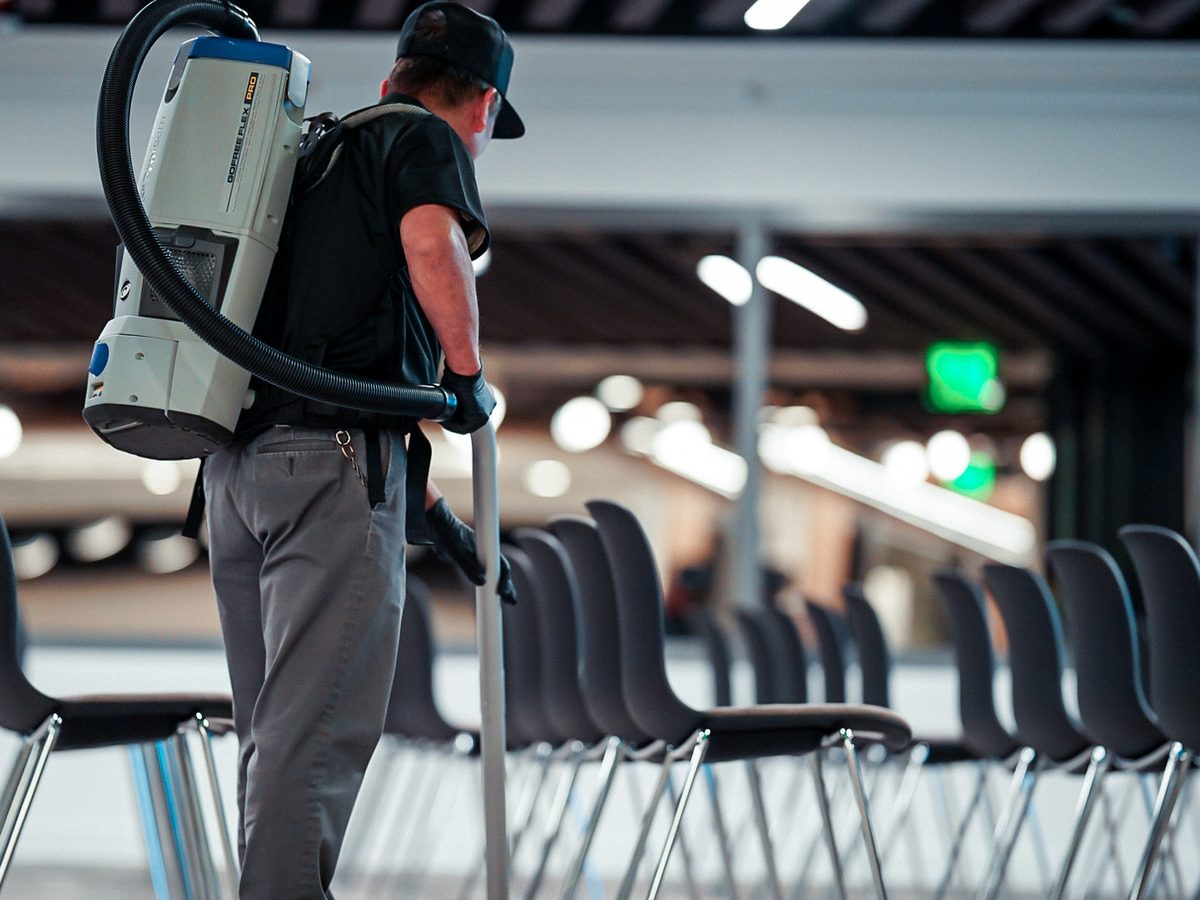 PBS crew member wearing a backpack vacuum cleaning between rows of chairs in a large open room.