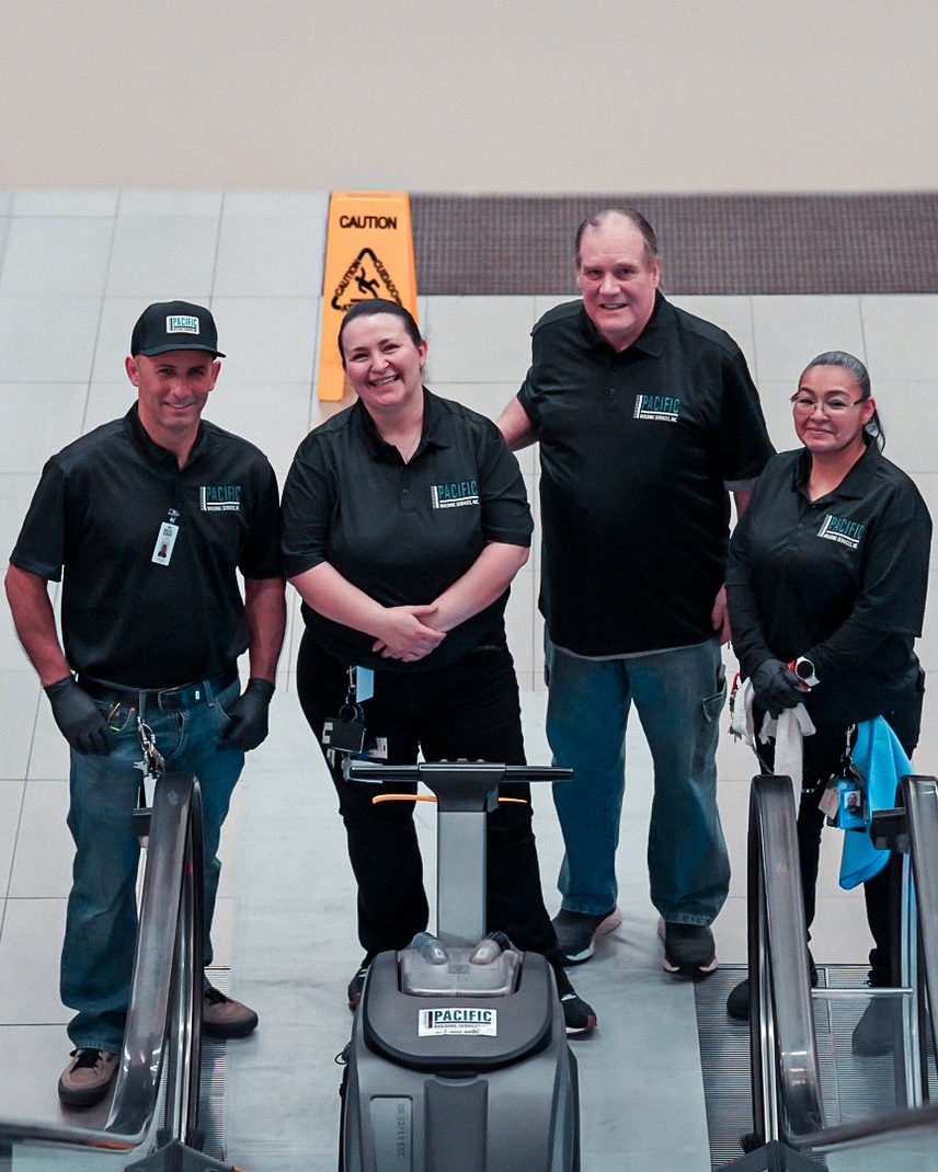 Four smiling PBS crew members standing with a floor scrubber at the top of an escalator, with wet-floor signs behind them.
