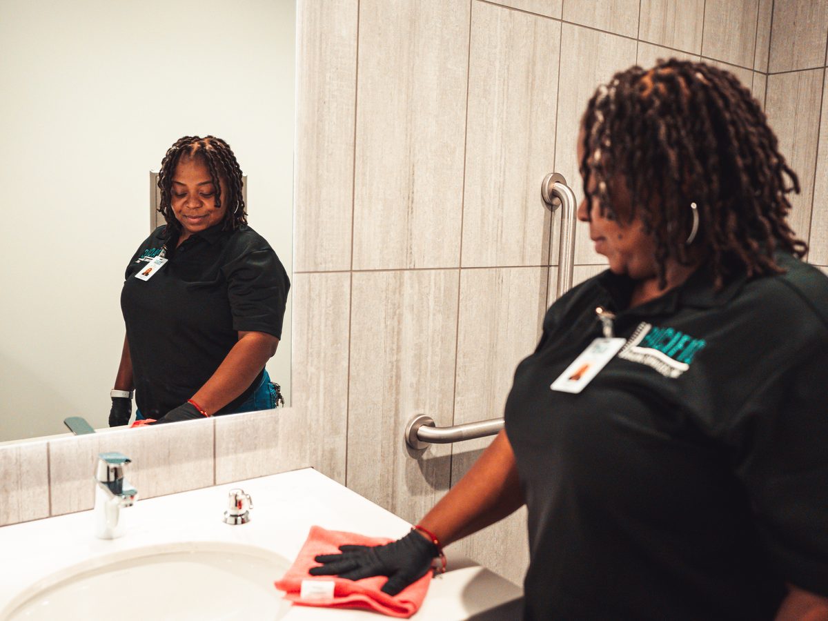 PBS crew member in gloves wiping a restroom sink counter with a red microfiber cloth, reflected in the mirror.