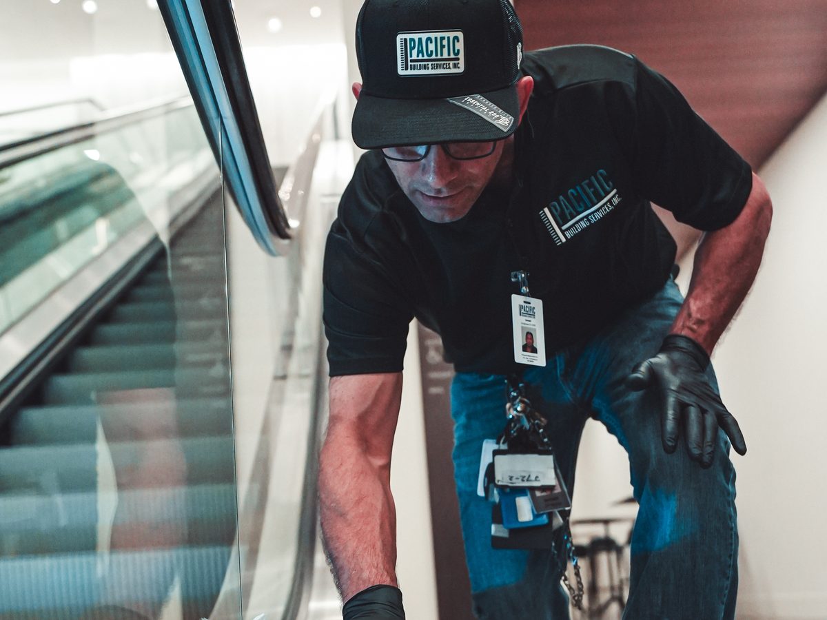 PBS crew member in gloves and glasses leaning down to detail-clean an escalator.