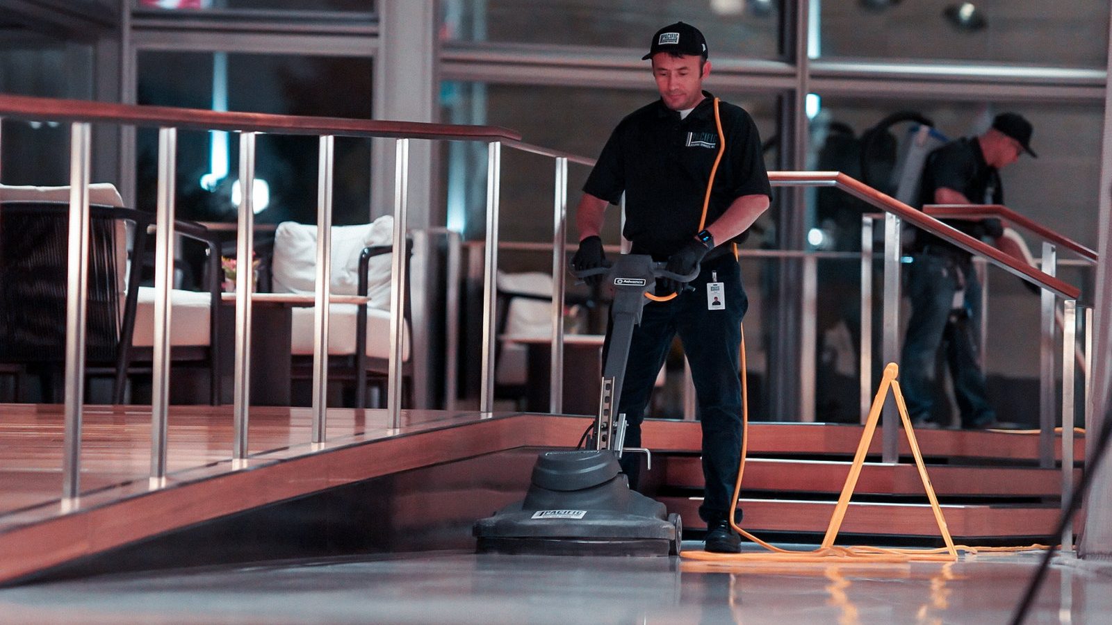 PBS floor technician in a black cap and polo running a floor burnisher across a polished lobby floor beside a curved stair railing, with a crew member wearing a backpack vacuum in the background.