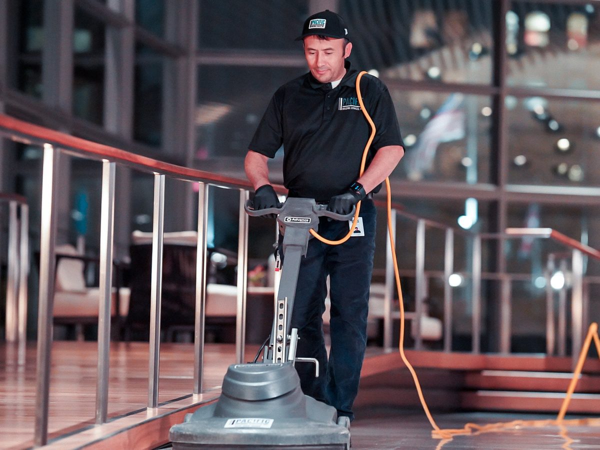 PBS floor technician in gloves running a floor burnisher across a polished lobby floor beside a curved railing.