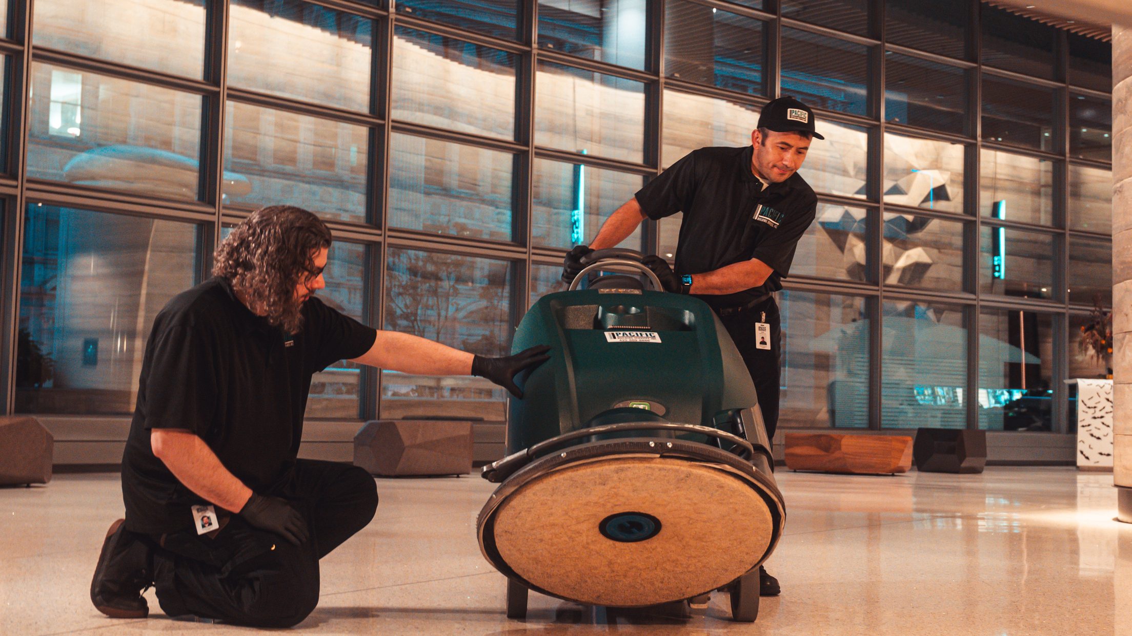 PBS floor technician kneeling to check a floor machine while a crew member steers it across a lobby lined with glass windows.