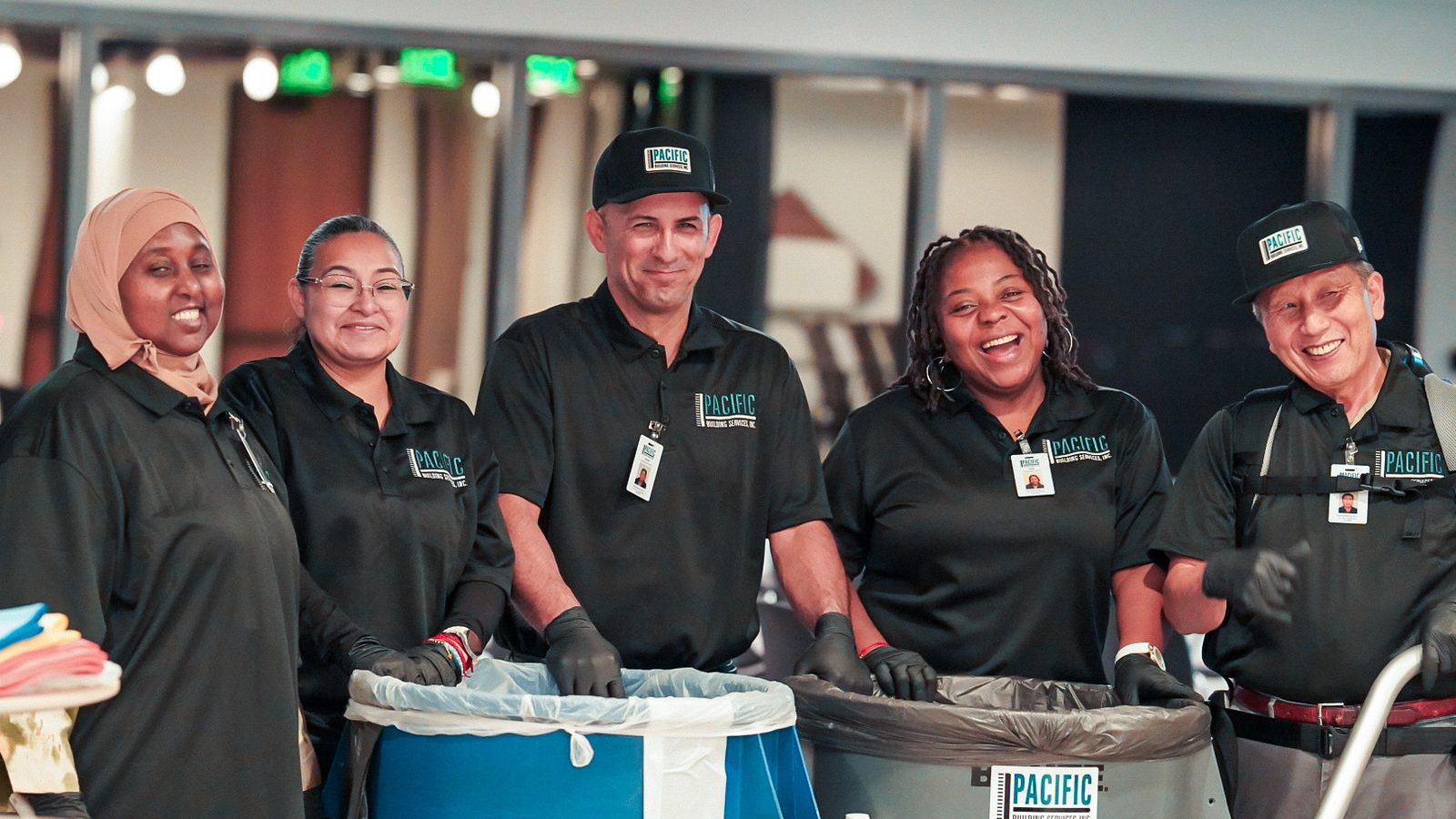 Five PBS crew members in gloves and black uniforms standing behind janitorial carts in a bright open space.
