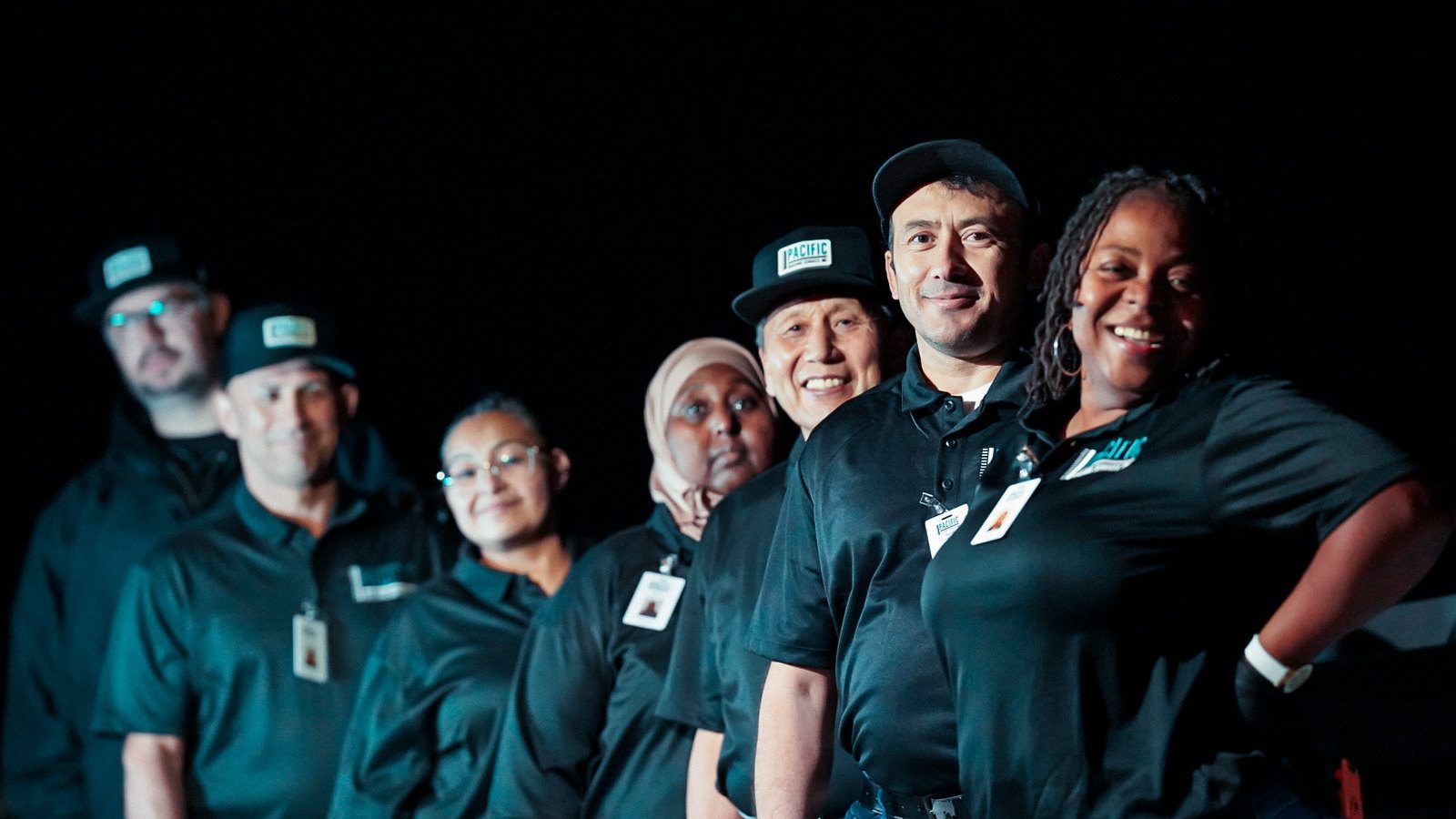 Seven PBS crew members in black uniforms and caps lined up and smiling against a dark background.
