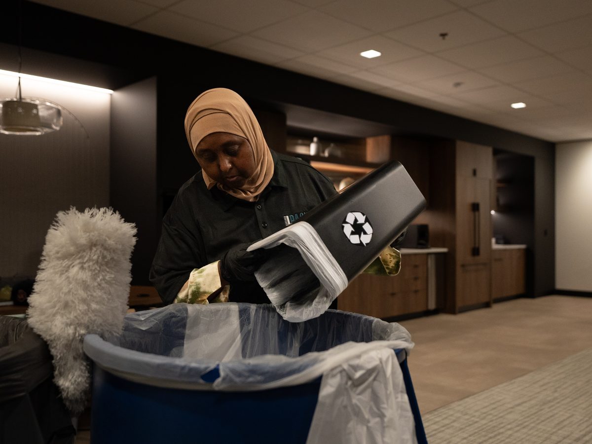PBS janitor in gloves and a headscarf emptying a bin into a blue recycling cart in an office corridor.