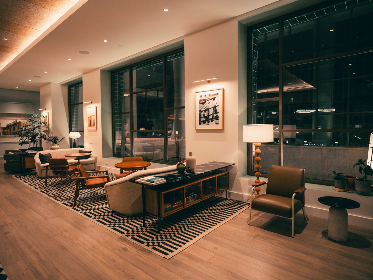 Lit office building lobby lounge at night with armchairs, a patterned rug, wall art and tall windows.
