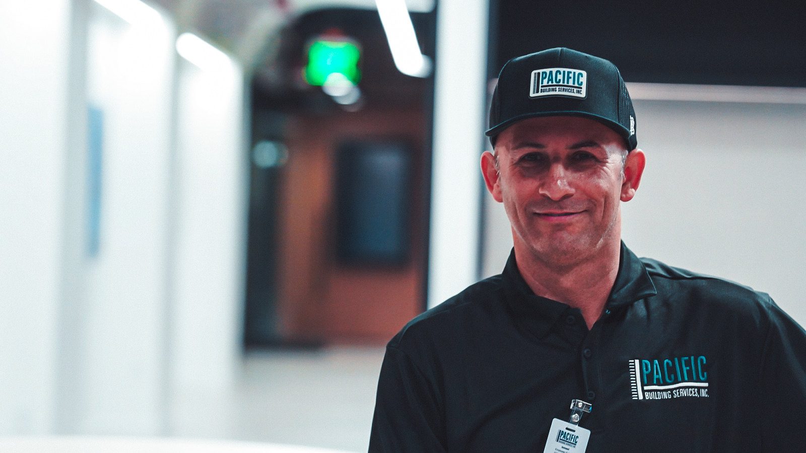 Smiling PBS crew member in a black PBS cap, polo and ID badge in an office.