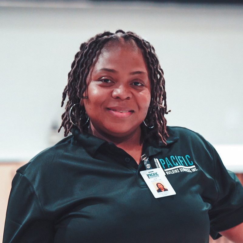 Smiling PBS crew member with shoulder-length locs, wearing a black PBS polo and ID badge.