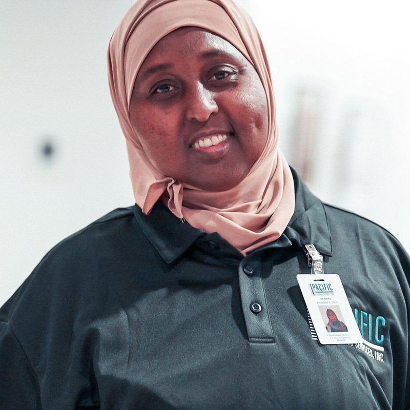 Smiling PBS crew member in a tan headscarf and a black PBS polo with ID badge.