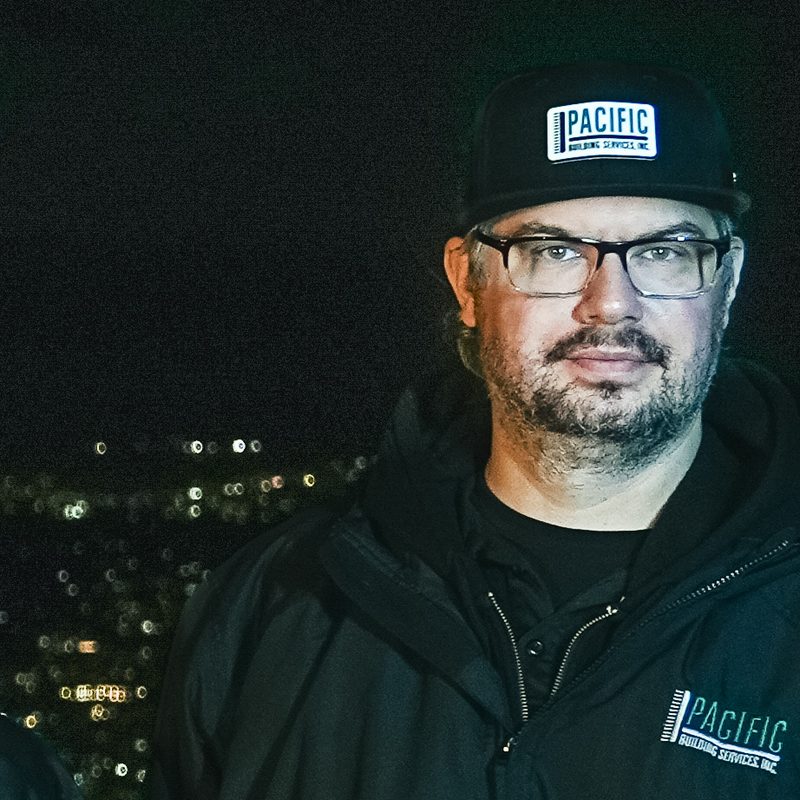 PBS crew member with glasses and a short beard, wearing a black PBS cap and PBS zip jacket, with city lights behind.