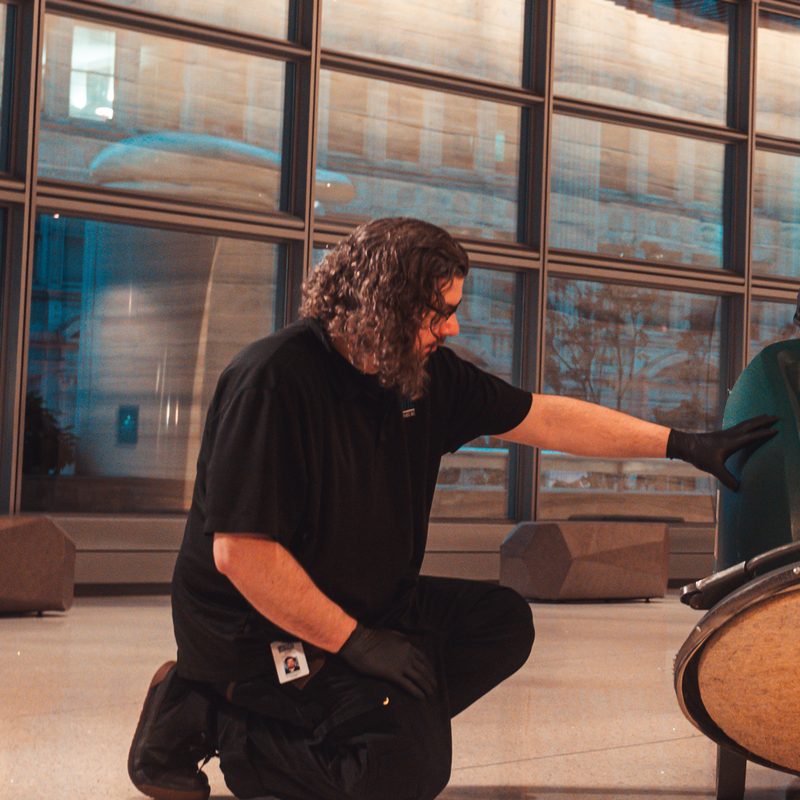 PBS floor technician with long curly hair, kneeling beside a floor machine in gloves.