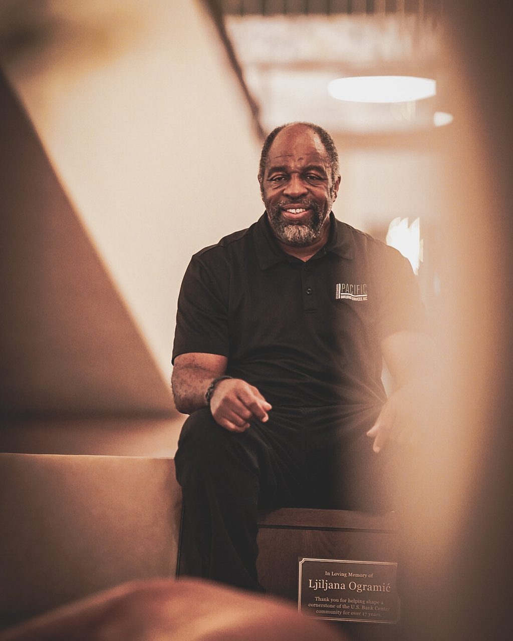 Smiling PBS team member in a black PBS polo seated on a lobby bench, with a plaque on the bench reading 